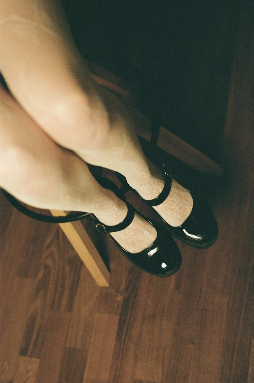 Füße in weißen Strumpfhosen mit schwarzen Lackschuhen, fotografiert von oben, auf Holzboden stehend.