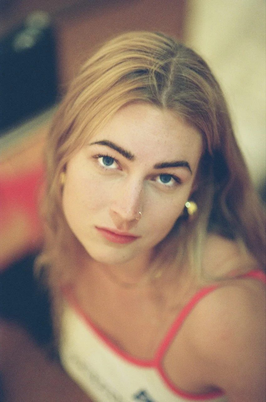 Nahaufnahme eines jungen Mädchens mit blonden Haaren, blauen Augen, Nasenpiercing und roten Trägern eines Tops, schaut in die Kamera.