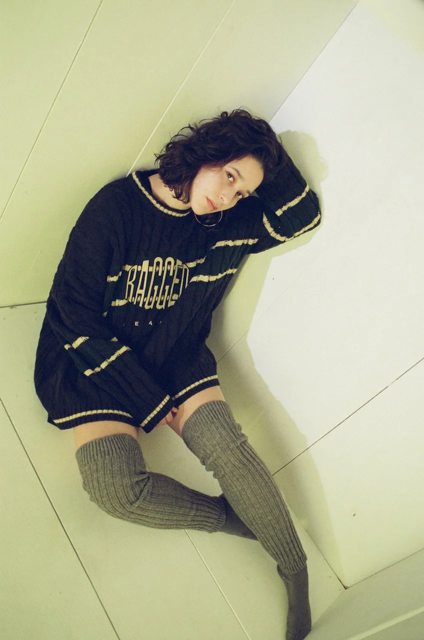 Junge Frau mit schwarzen zerrissenen Pullover und grauen Strümpfen sitzt an der Ecke einer weißen Wand und blickt in die Kamera.