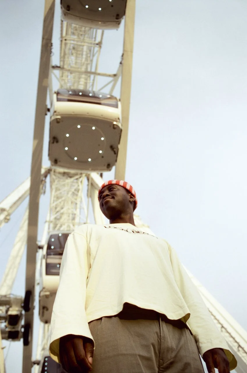 Junge Person vor dem Riesenrad, blickt nach oben, trägt gelbes langärmeliges Oberteil und rote Mütze, Himmel im Hintergrund.