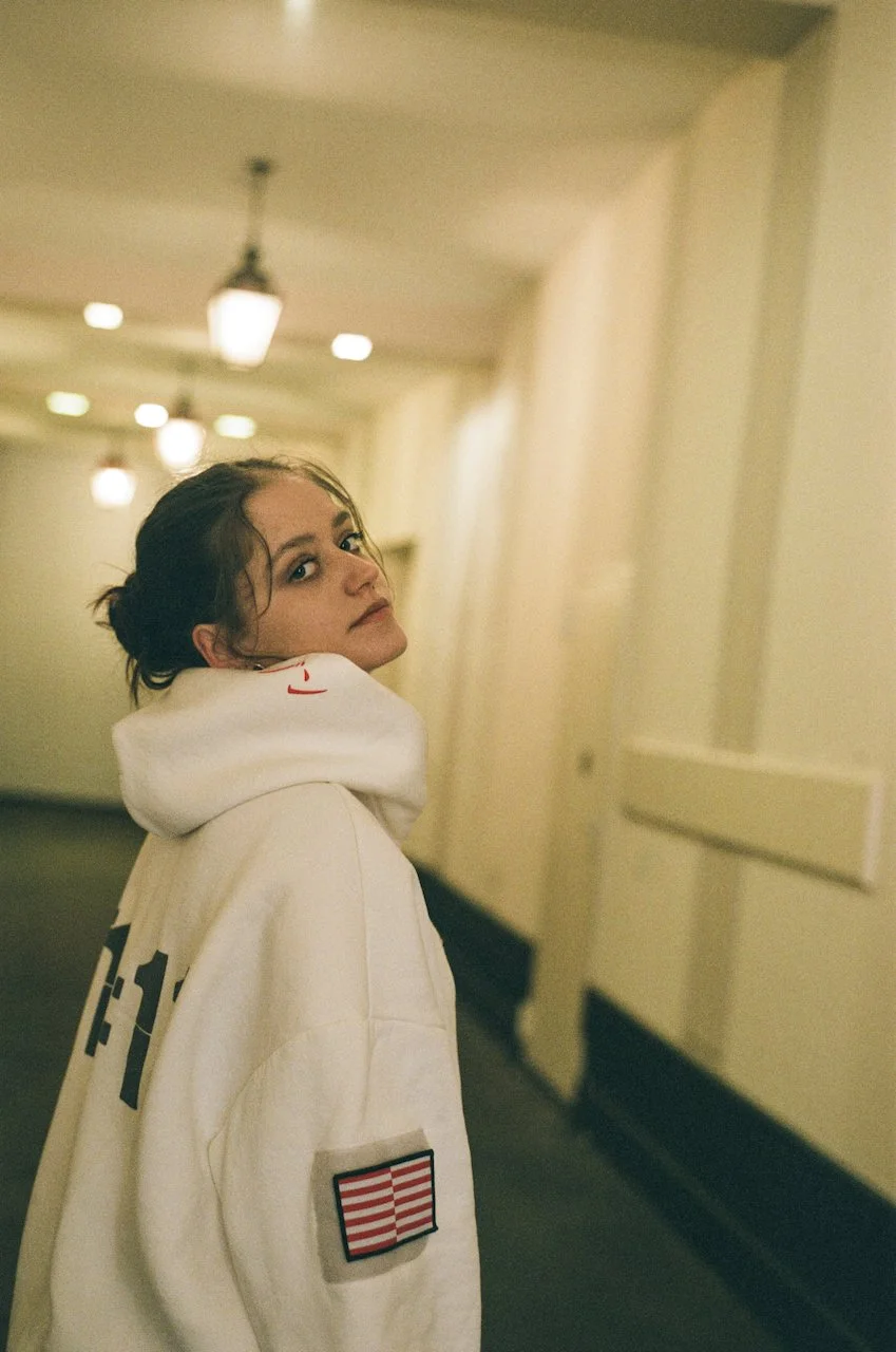 Junge Frau mit dunklem Haar, die eine weiße Kapuzenjacke trägt, in einem Flur mit warmem Licht und Deckenleuchten.