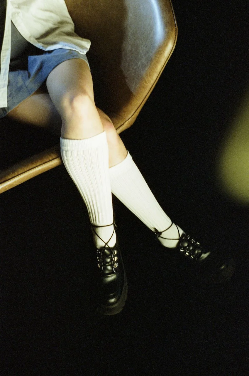 Das Bild zeigt die Beine einer Person, die auf einem braunen Sitz liegt, mit einem grauen Hemd, weißen hohen Socken und schwarzen Schuhen mit Schnürung.