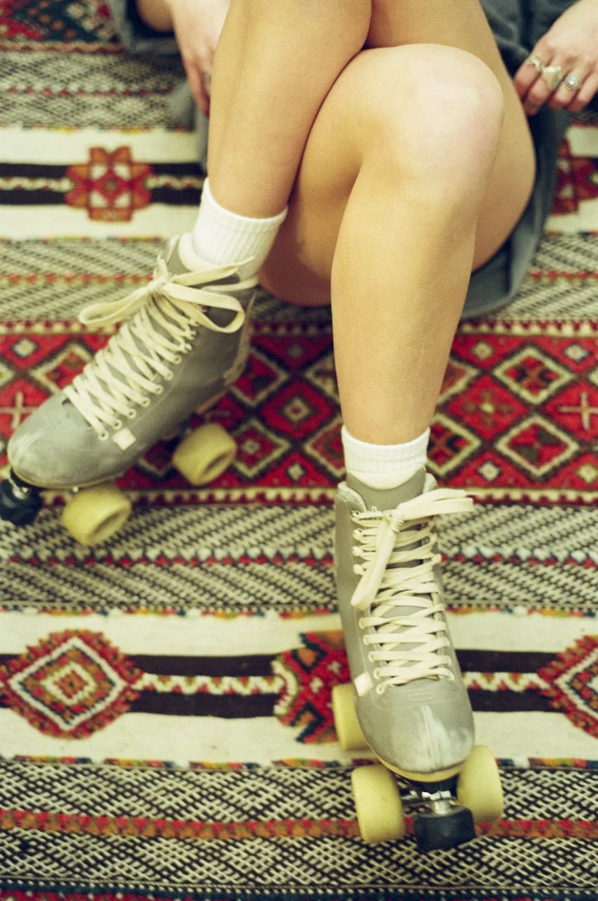 Person sitzt auf einem Teppich und trägt Rollschuhe mit weißen Schnürsenkeln, weiße Socken und eine kurze graue Shorts.