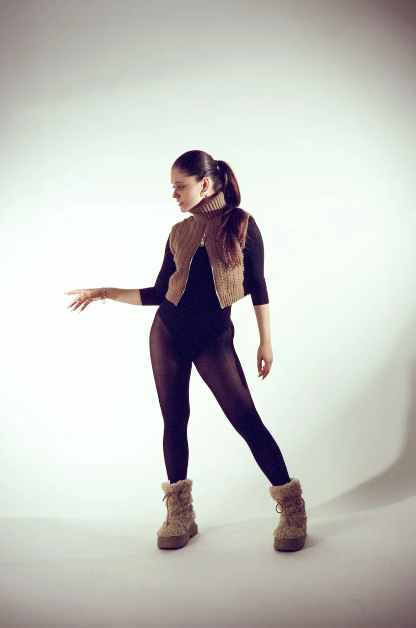 Frau trägt schwarze Leggings, eine braune Strickweste und flauschige beige Stiefel, posiert vor weißem Hintergrund.