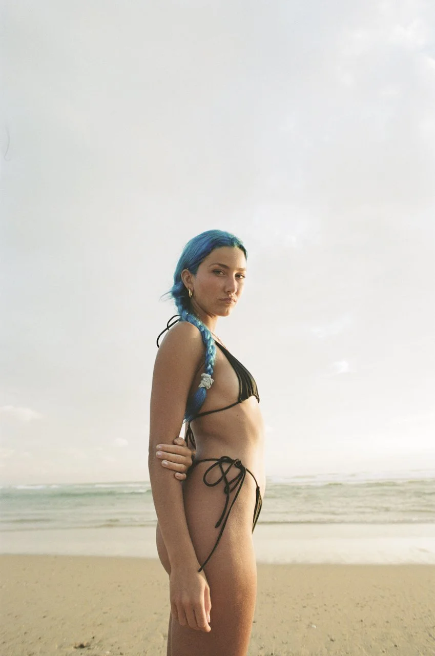 Junge Frau im Bikini mit blauer Zopffrisur am Strand, Blick schräg nach vorne