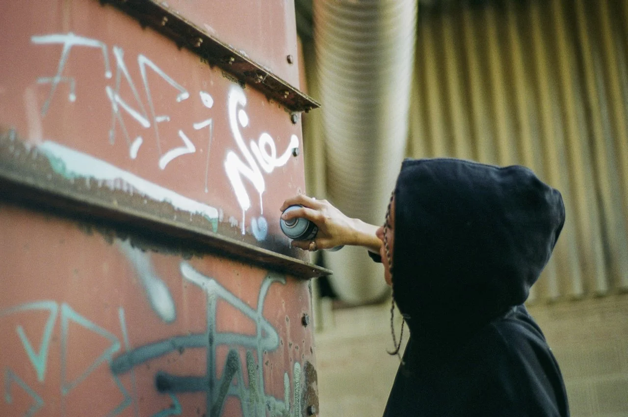 Junge trägt schwarze Kapuzenjacke, sprüht Graffiti auf eine Wand in einem Raum mit grünen Vorhängen.