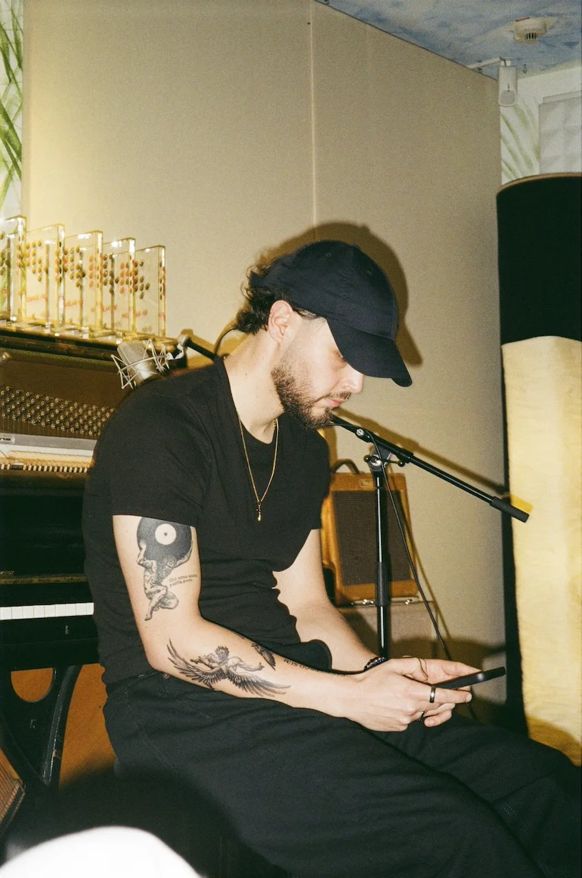 Junger Mann in schwarzem T-Shirt und schwarzem Cap sitzt in einem Raum mit einem Klavier im Hintergrund, schaut auf sein Smartphone.