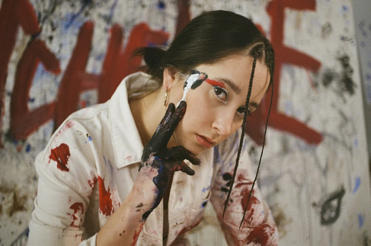 Junge Frau klebt bei einem Kunstprojekt an einer Wand mit roter, blauer und schwarzer Farbe, trägt weiße Jacke mit Farbflecken, hat dunkle Haare, trägt roten Augen-Make-up und goldene Ohrringe.