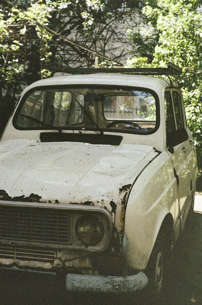 Ein alter, verrosteter weiße Auto steht vor einem Laubbaum, die Scheiben sind verschmutzt, und das Auto sieht vernachlässigt aus.