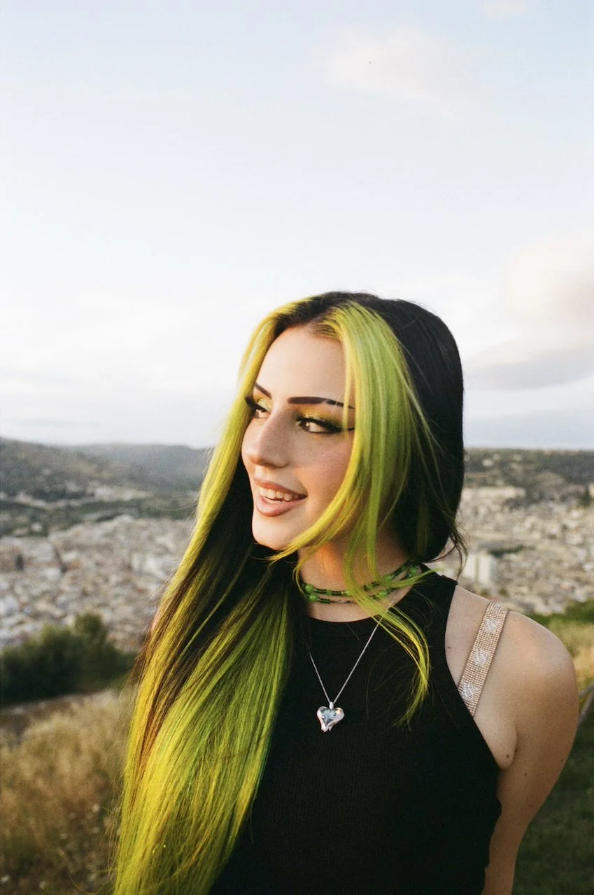 Junge Frau mit schwarz-grünen Haaren, lächelnd im Freien, mit Stadtlandschaft im Hintergrund.