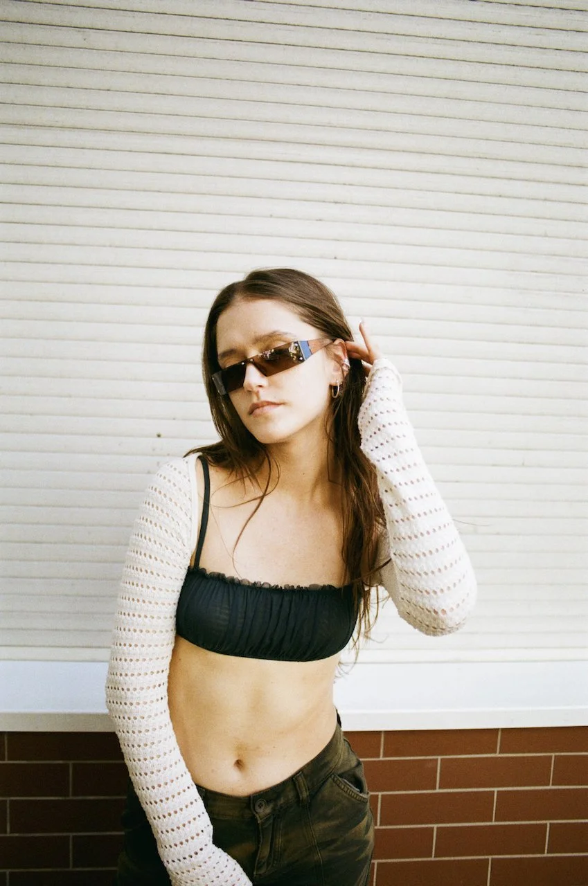 Junge Frau mit Sonnenbrille, schwarz Crop Top und weißen Hacken-Armlehn-Ärmeln steht vor einer weißen, horizontalen Lamellenwand, die an der unteren Seite von roten Ziegeln unterbrochen wird.