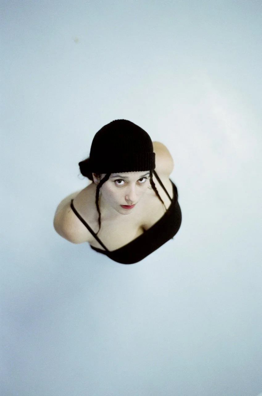 Junge Frau liegt auf dem Rücken auf dem Boden, trägt einen schwarzen Hut und ein schwarzes Kleid mit dünnen Trägern, blickt nach oben.