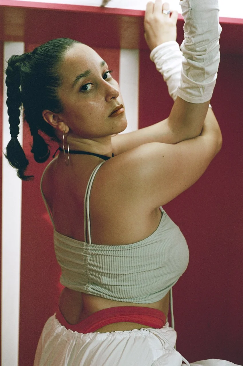 Junge Frau lehnt an einer roten Wand, schaut nach oben, trägt einen grauen Crop-Top, weiße Shorts, schwarze Halskette und große Ohrringe, mit einem lockeren weißen Tuch um den Arm.