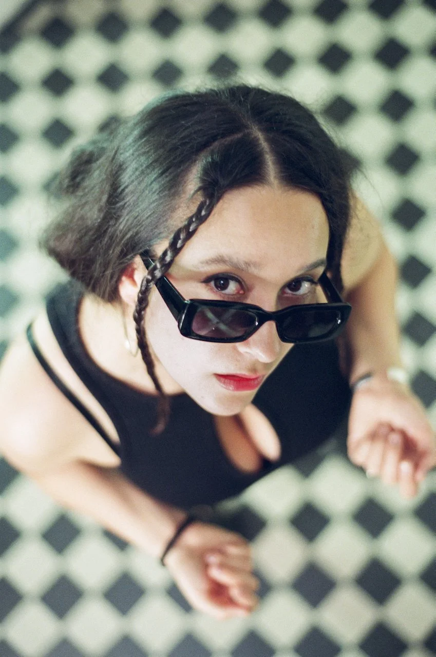 Frau mit dunklen Haaren und schwarzen Sonnenbrillen, Blick nach oben, auf einem Schwarz-Weiß-Fliesenboden fotografiert.