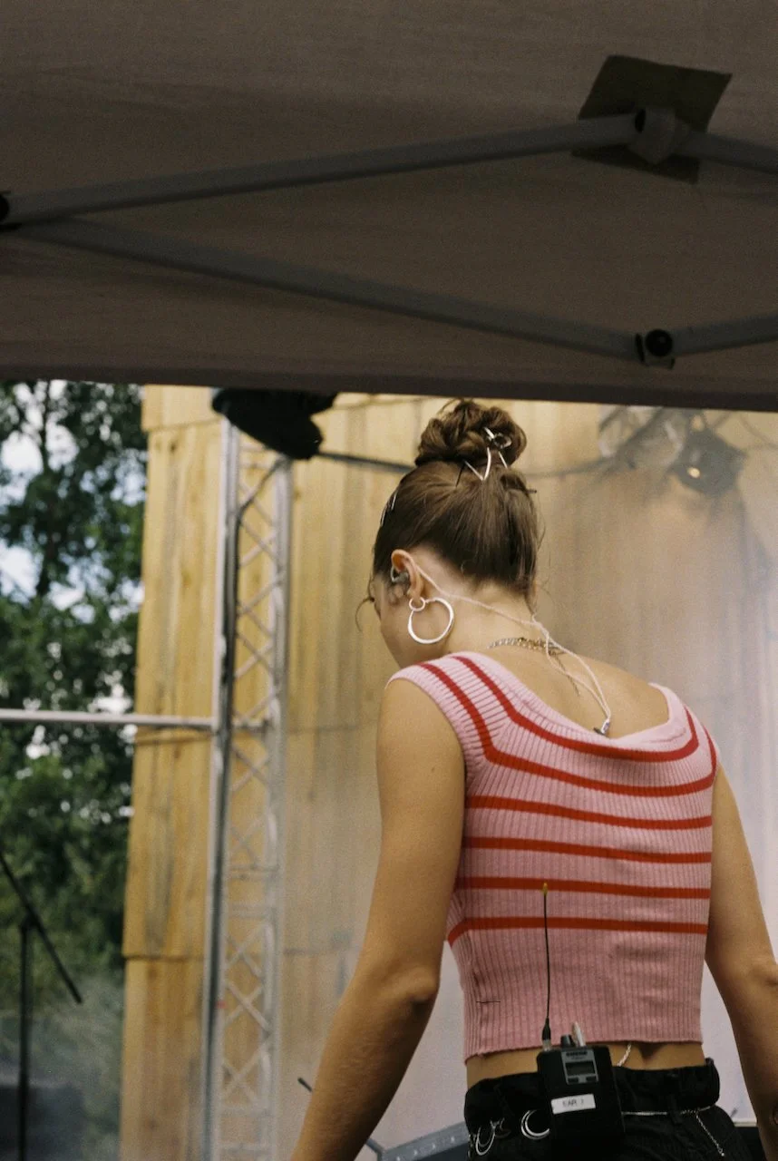 Eine junge Frau, die auf einer Bühne steht, trägt ein pink-rotes gestreiftes ärmelloses Top, hat ihre Haare hochgesteckt, trägt große Ohrringe und einen Kopfhörer sowie ein Mikrofon und eine Funk-Box auf der Taille.