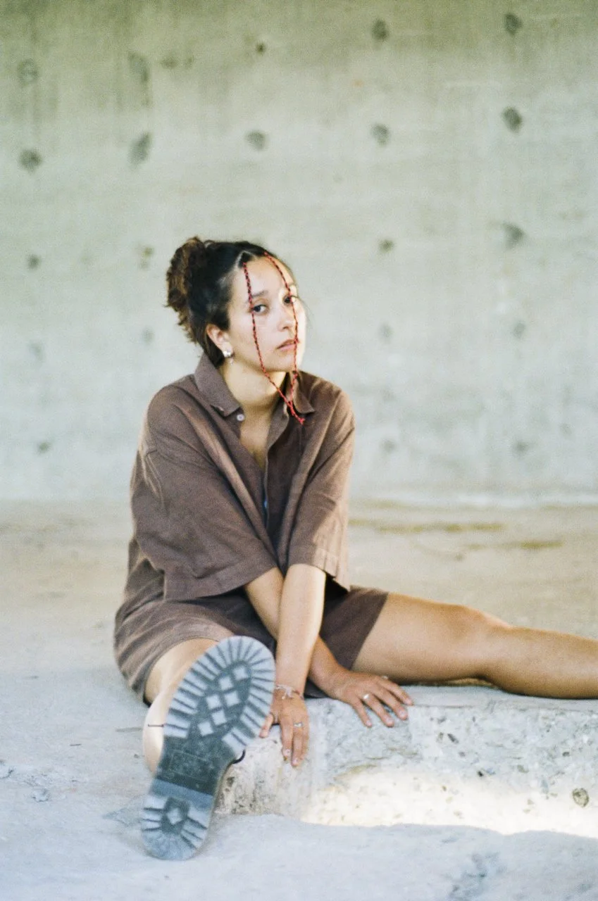 Junge Frau sitzt auf dem Boden, trägt einen braunen Hemd und Shorts, hat zerrissene Haare, trägt Ohrringe und Schmuck, in einem Raum mit Betonwand, nüchternem Ambiente.
