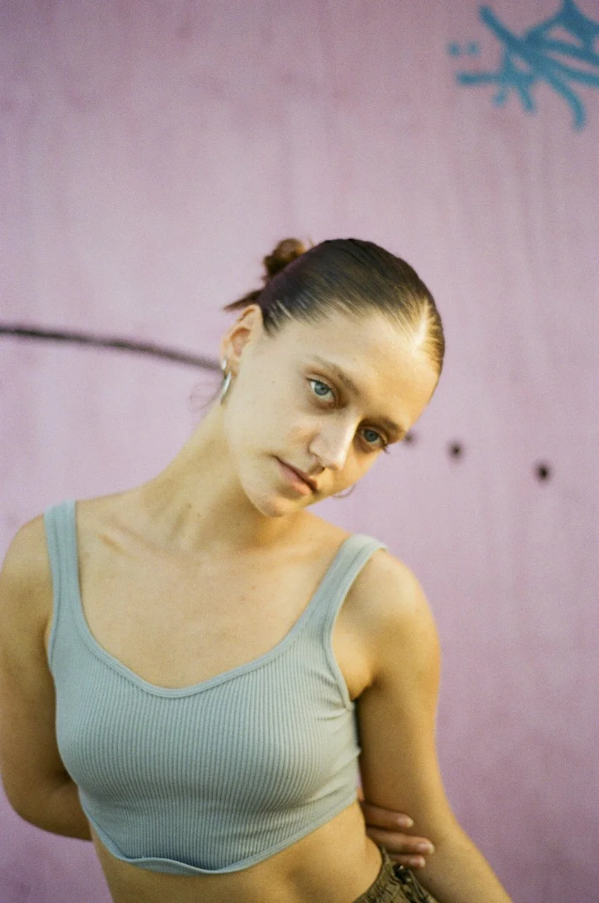 Junge Frau mit blauen Augen und dunklem Haar, zusammengebunden, trägt ein hellblaues Top, steht vor einer pinken Wand mit Graffiti.