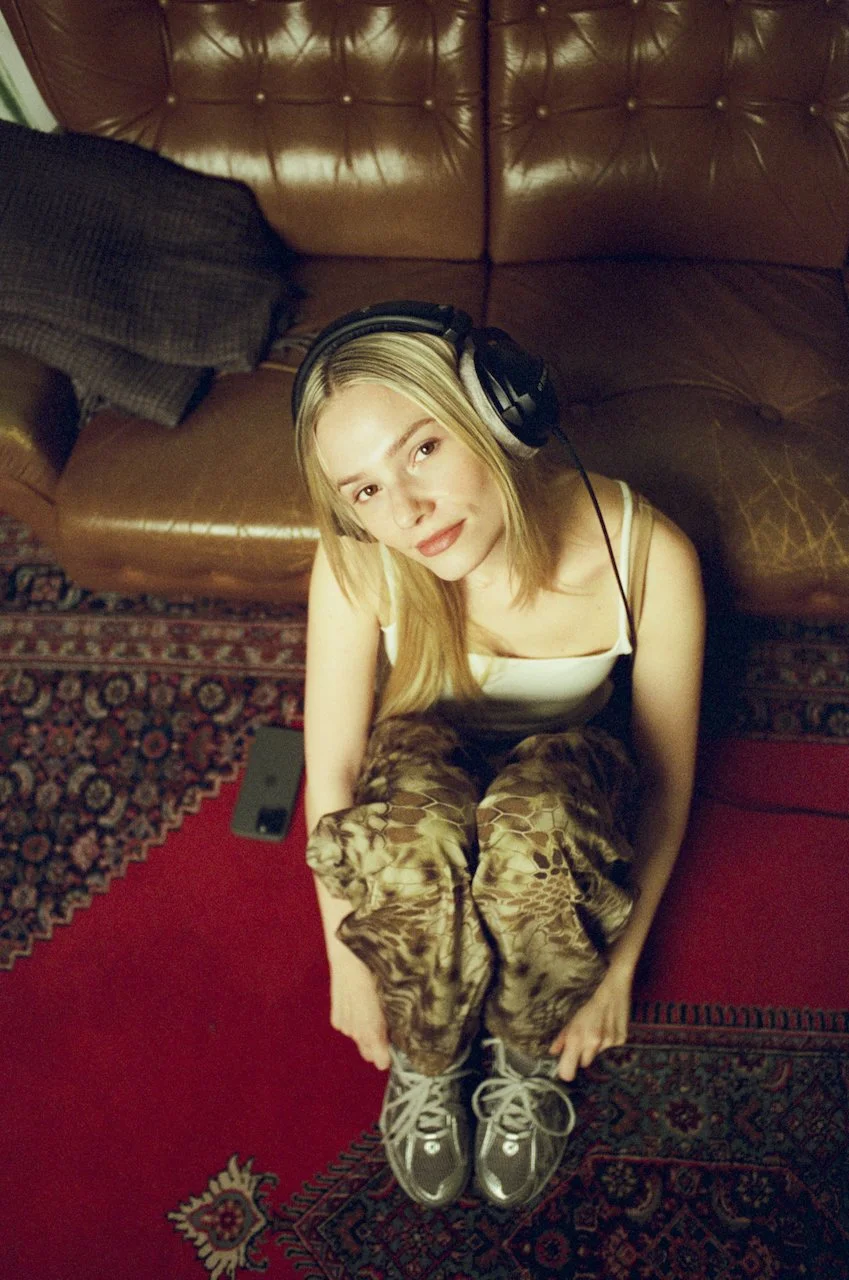 Junge Frau mit Kopfhörern, sitzend auf einem roten Teppich, blickt nach oben, trägt kariertes Kleid, braune Sneakers, neben braunem Ledersofa.