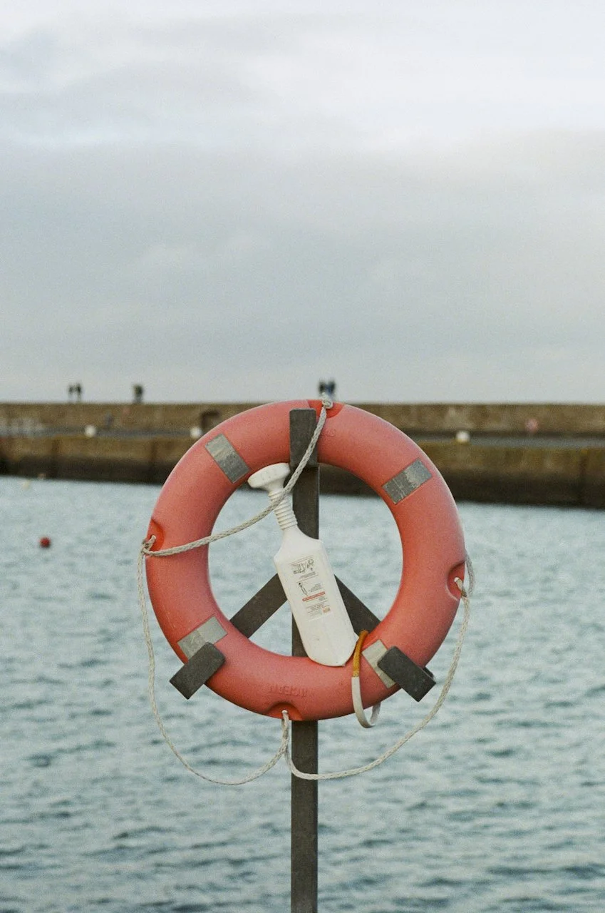 Rettungsring mit Rettungsflagge, an einem Pfosten am Wasser befestigt, im Hintergrund eine Hafenkulisse bei bewölktem Himmel.