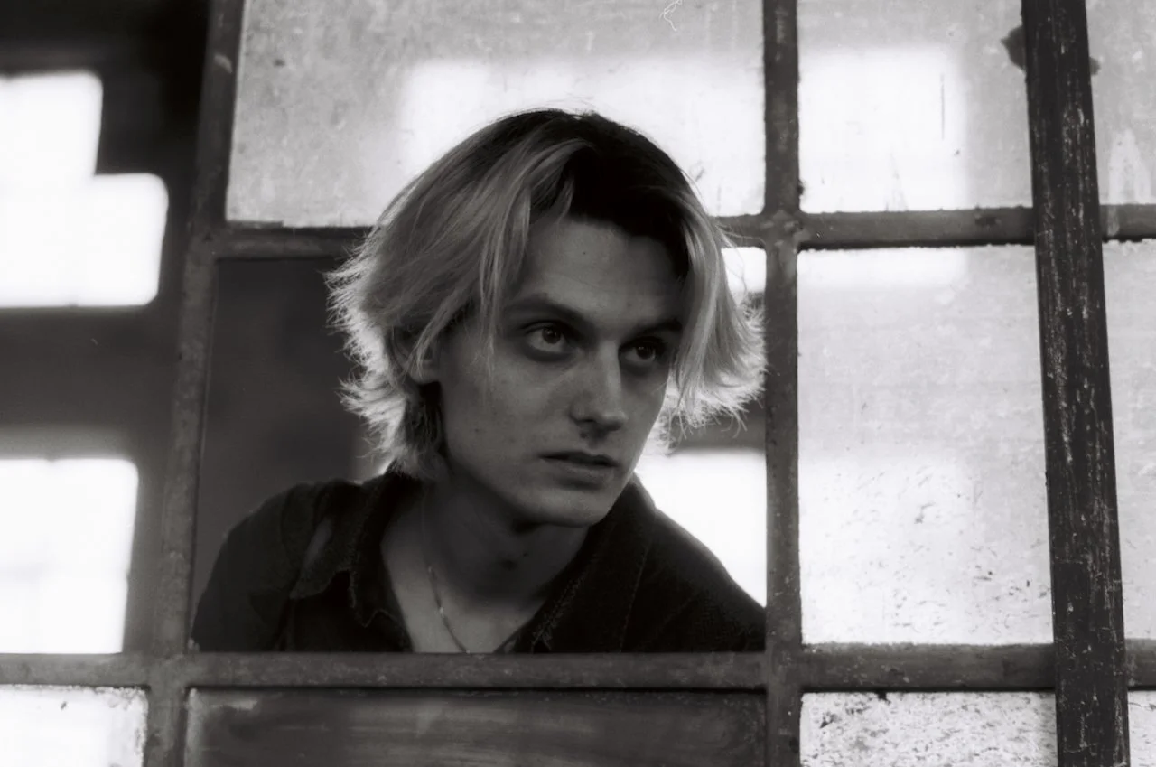 Schwarz-weiß-Foto eines jungen Mannes mit blondem, unordentlichem Haar blickt durch ein verrostetes Gitterfenster.