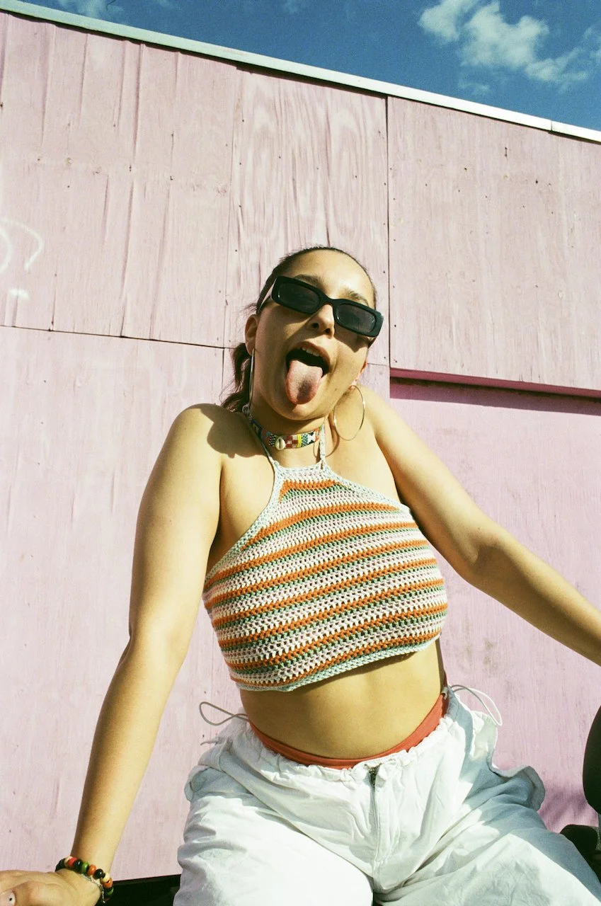 Junge Frau mit Sonnenbrille und buntem Stricktop, die die Zunge ausstreckt, vor einer pinken Wand und blauem Himmel.