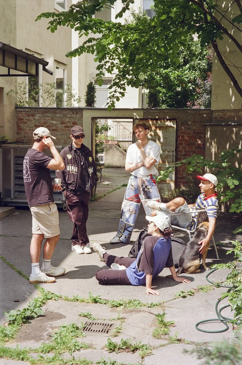 Fünf junge Menschen in einem urbanen Innenhof, einer sitzt auf dem Boden, die anderen stehen, darunter ein junger Mann mit einem Hund, alle scheinen im Gespräch zu sein. Es ist sonnig und es gibt Grünpflanzen.