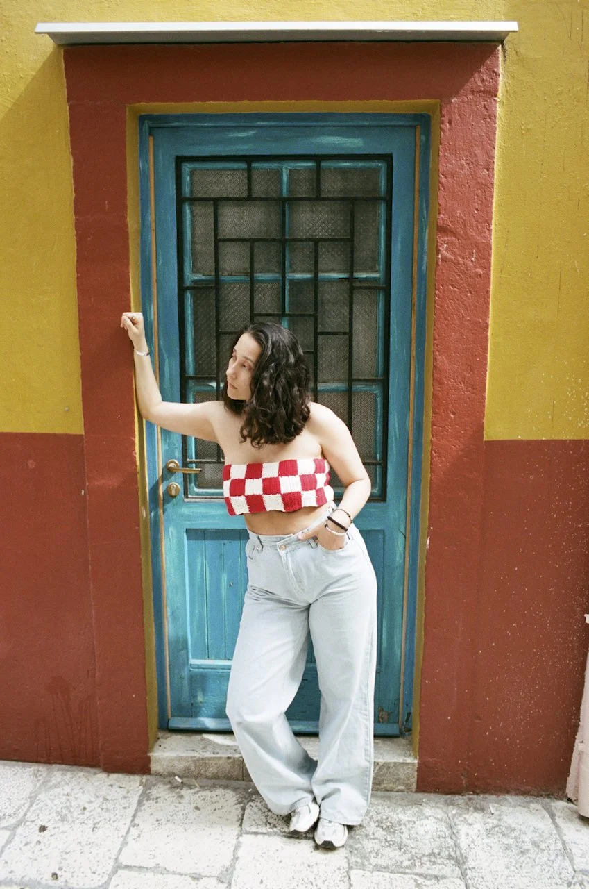 Junge Frau mit lockigem schwarzen Haar, trägt eine rote und weiße Karotten-Bustier, helle Jeans, steht vor einer bunten Tür, die umrahmt ist von hellgelber und rotbrauner Wand, auf einem gepflasterten Boden.