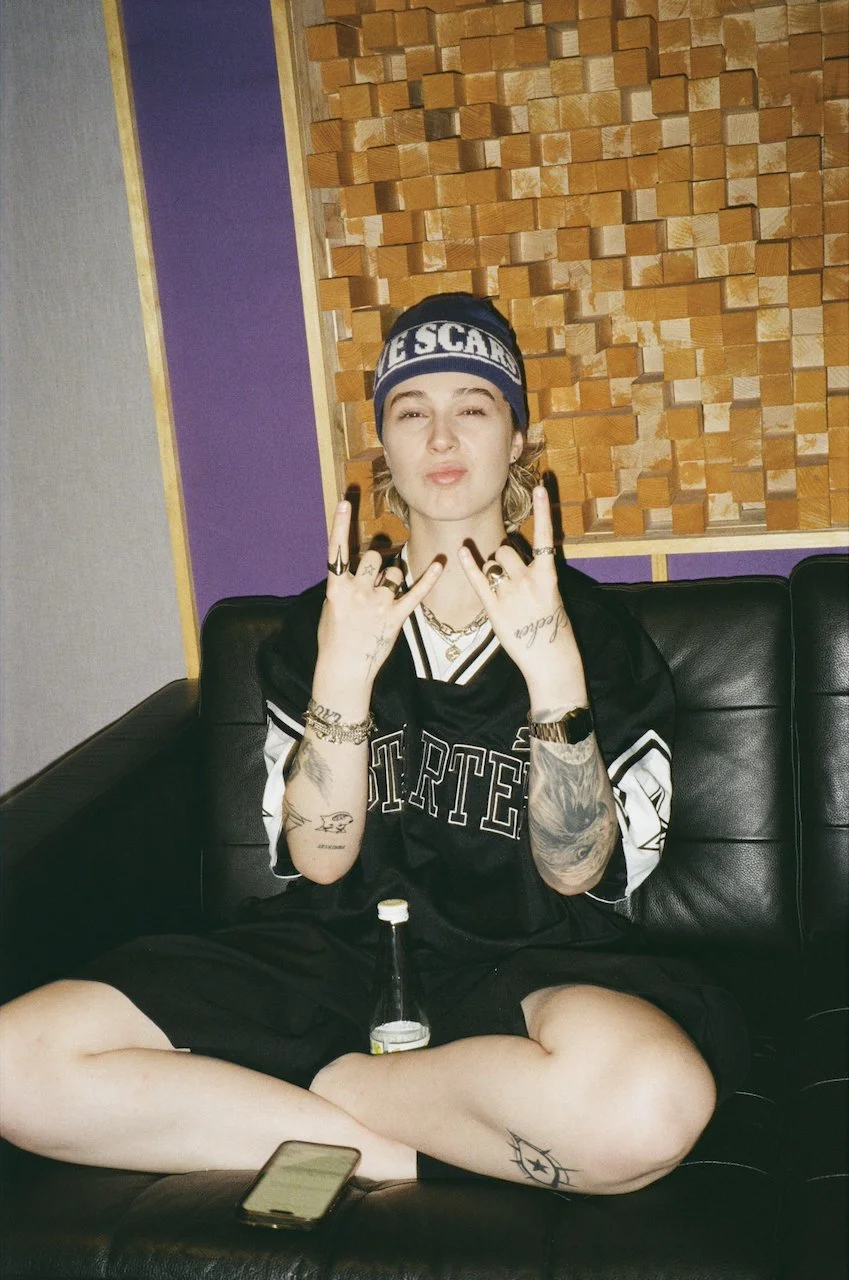 Junge sitzt auf einem schwarzen Sofa, trägt eine schwarze Sportjacke und eine Baskenmütze, macht Handzeichen, hat Tattoos an Armen und Beinen, hält eine Flasche und hat ein Smartphone vor sich.