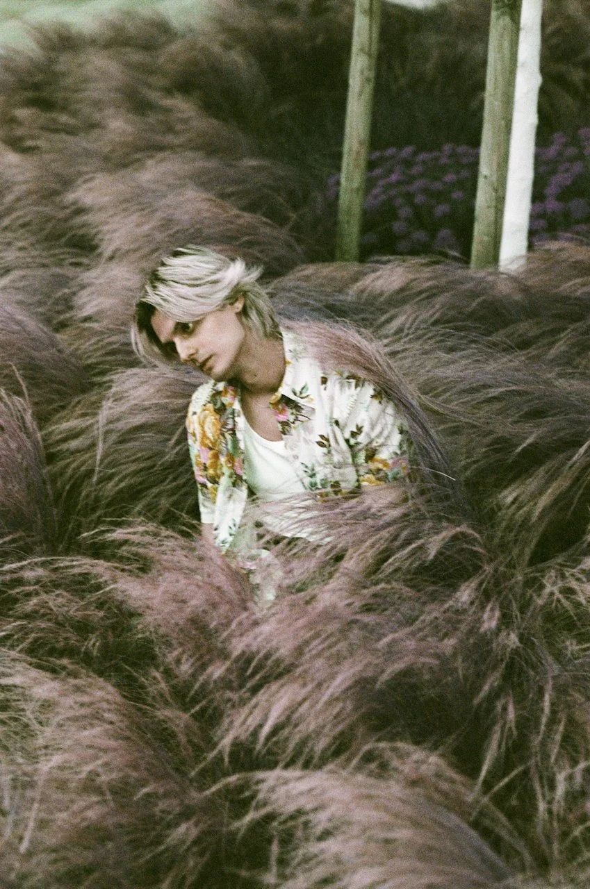 Ein junger Mensch mit blond-schwarzhaarigem Haar sitzt inmitten einer Gruppe von brauen, langen Haaren, in einer Wiese mit Holzpfählen im Hintergrund.