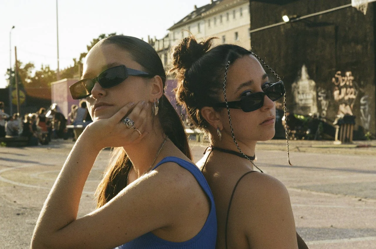 Zwei junge Frauen mit dunklen Sonnenbrillen stehen Rücken an Rücken in einer Stadt im Sonnenlicht, umgeben von Menschen und Graffiti-Wänden.
