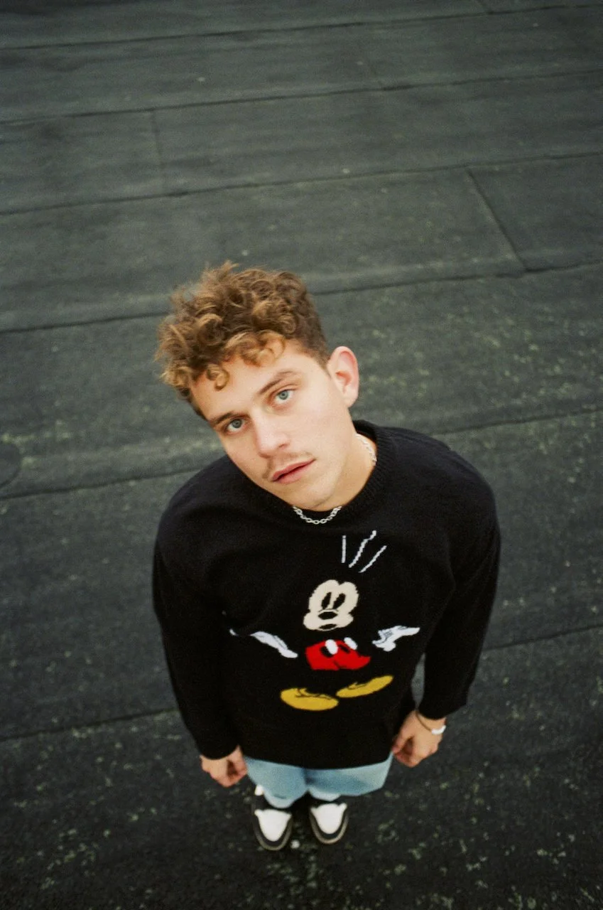 Junge mit lockigem Haar trägt einen schwarzen Mickey Mouse Pullover und steht auf einer dunklen Asphaltstraße, Blick nach oben gerichtet.