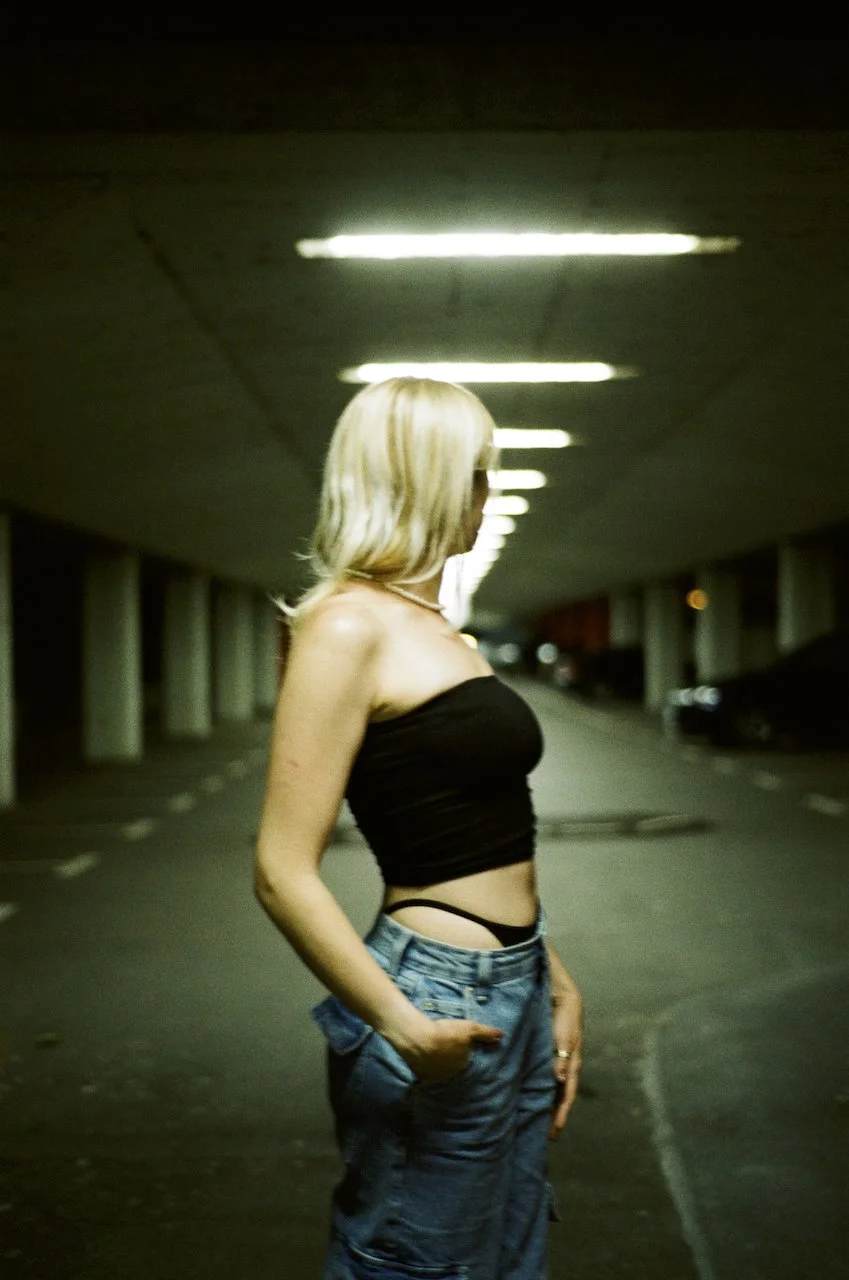 Junge Frau mit blonder trage-lose Oberteil und Jeans in einer dunklen, unterirdischen Parkgarage bei Nacht.