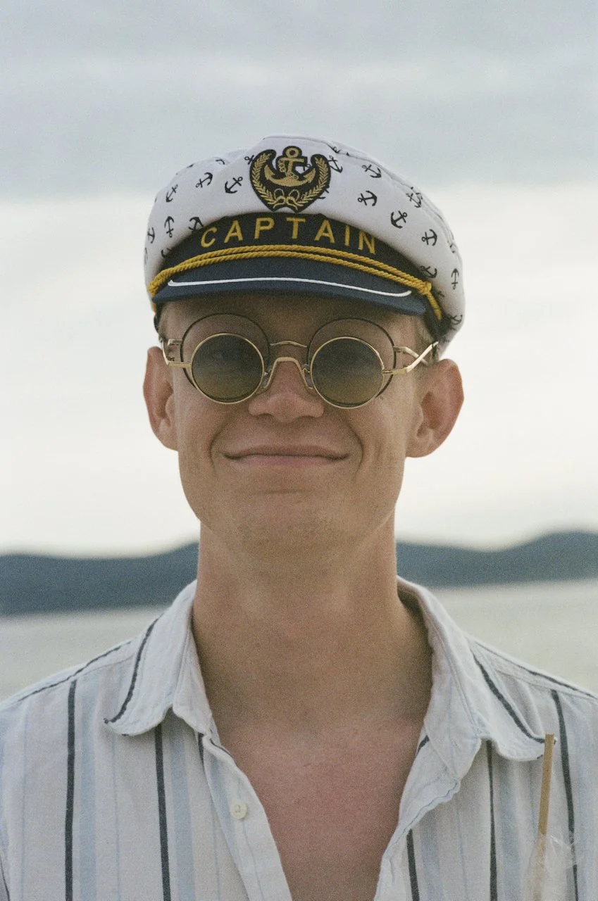 Ein Mann trägt eine weiße Matrosenmütze mit Anker- und Affen-Symbolen, einer goldenen Borte mit der Aufschrift »CAPTAIN«, sowie eine Brille mit runden schwarzen Gläsern