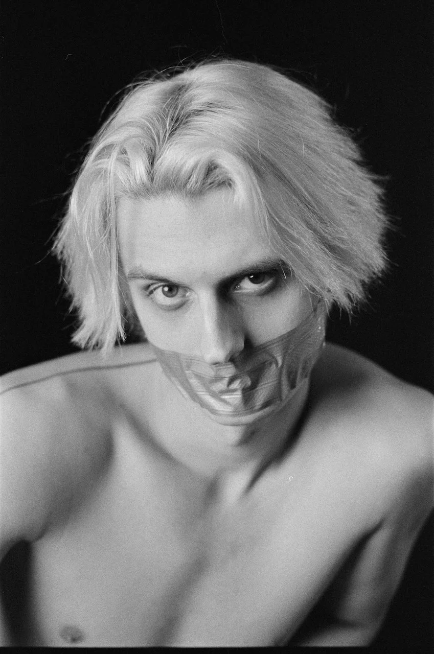 Schwarze und weiße Fotografie eines nackten Mannes mit maskiertem Mund, Blick direkt in die Kamera, mit blondem, schulterlangem Haar vor dunklem Hintergrund.