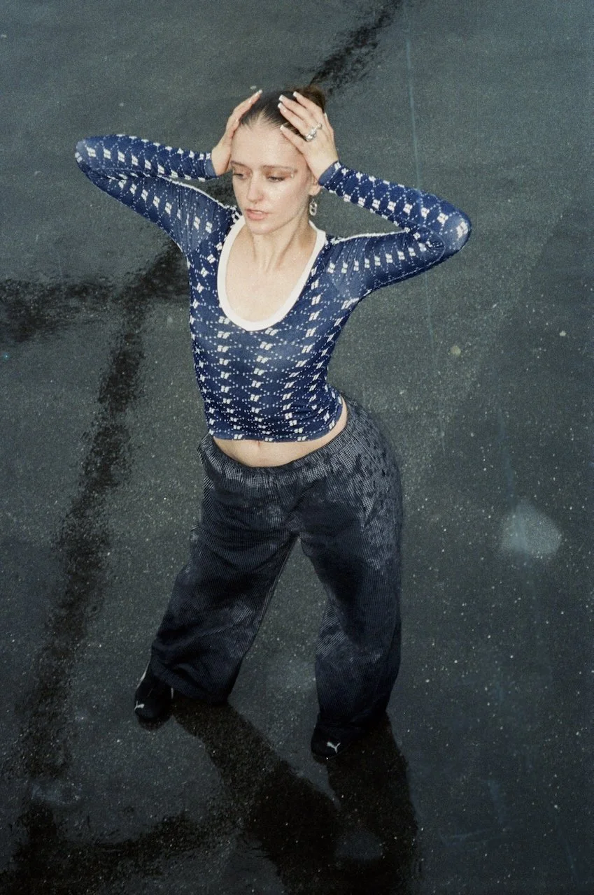 Eine junge Frau steht in einem Wasserbecken, trägt eine dunkelblaue, transparente, langärmlige Top und schwarze Sportschuhe, hält sich den Kopf mit beiden Händen und schaut nach unten.