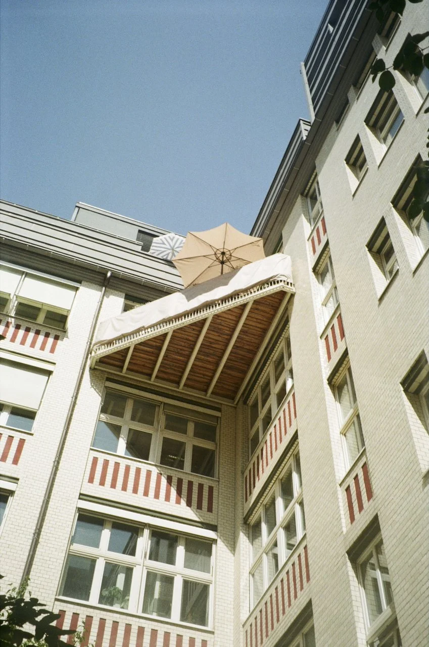 Blick nach oben auf einen Balkon mit Sonnenschirm vor einem modernen weißen Gebäudekomplex bei blauem Himmel.