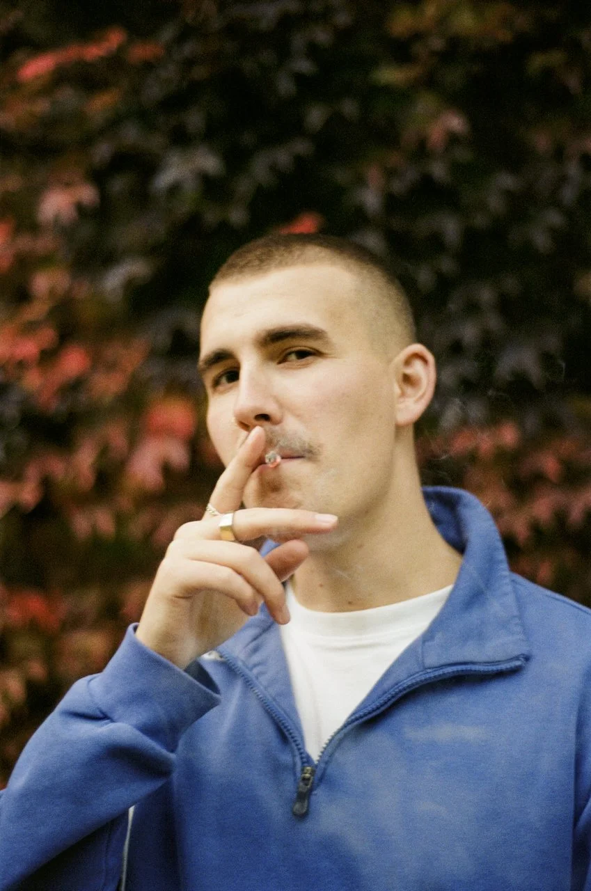 Junge Mann in blauer Jacke, der an einer Zigarette zieht, vor einem Hintergrund mit rot und dunkelgrünen Blättern.