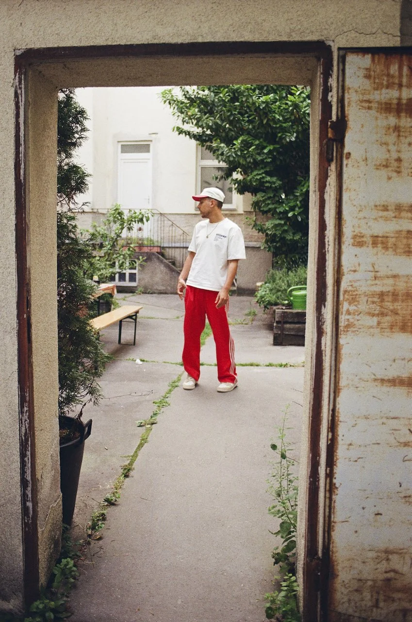 Ein Mann in weißen T-Shirt, roten Jogginghosen und einer weißen Kappe steht im Hinterhof, umgeben von Bäumen und Pflanzen.