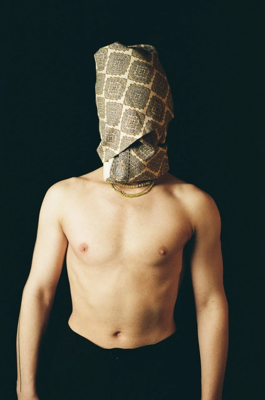 Ein nackter Mensch mit einem gestaltartigen Stoff über dem Kopf, der ein Muster mit geometrischen Formen trägt, vor einem schwarzen Hintergrund.