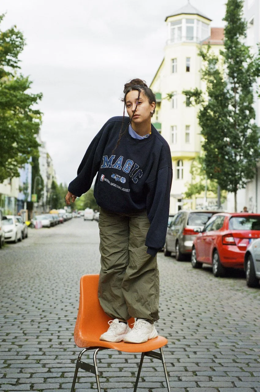 Junge steht auf einem orangefarbenen Stuhl auf einer Kopfsteinpflasterstraße, umgeben von Häusern und Bäumen.