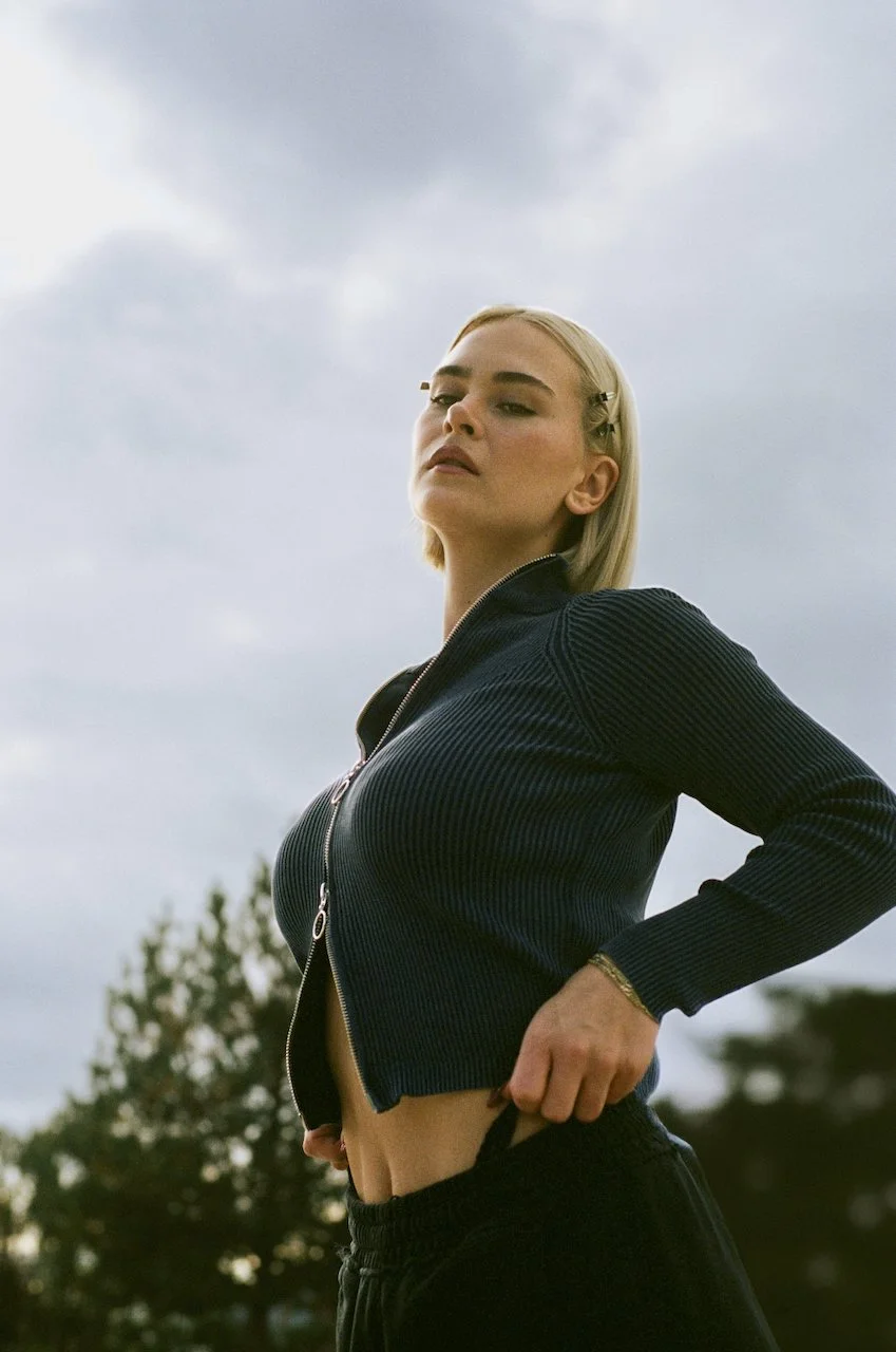 Junge Frau mit blonder Kurzhaarfrisur trägt schwarze Zipperjacke und schwarze Hose, steht im Freien mit Himmel und Bäumen im Hintergrund.