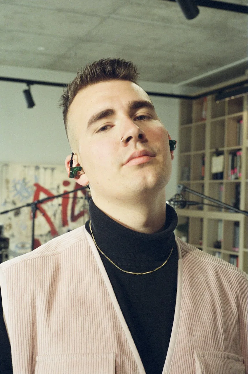 Junge mit Kurzhaarschnitt, Piercing, schwarze Kleidung, in einem Raum mit Bücherregalen und Graffiti im Hintergrund.