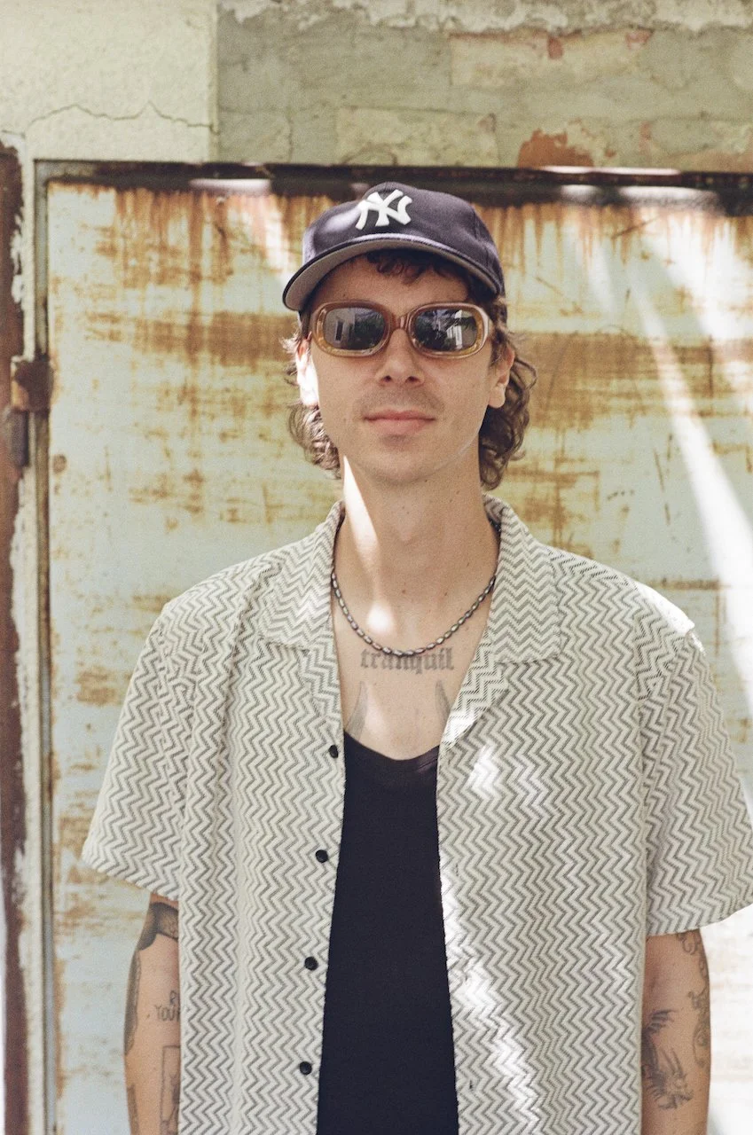 Junger Mann mit dunklem Cap, Sonnenbrille, kurzärmligem Hemd und Tattoos, vor einer alten, verrosteten Metalloberfläche im Hintergrund.