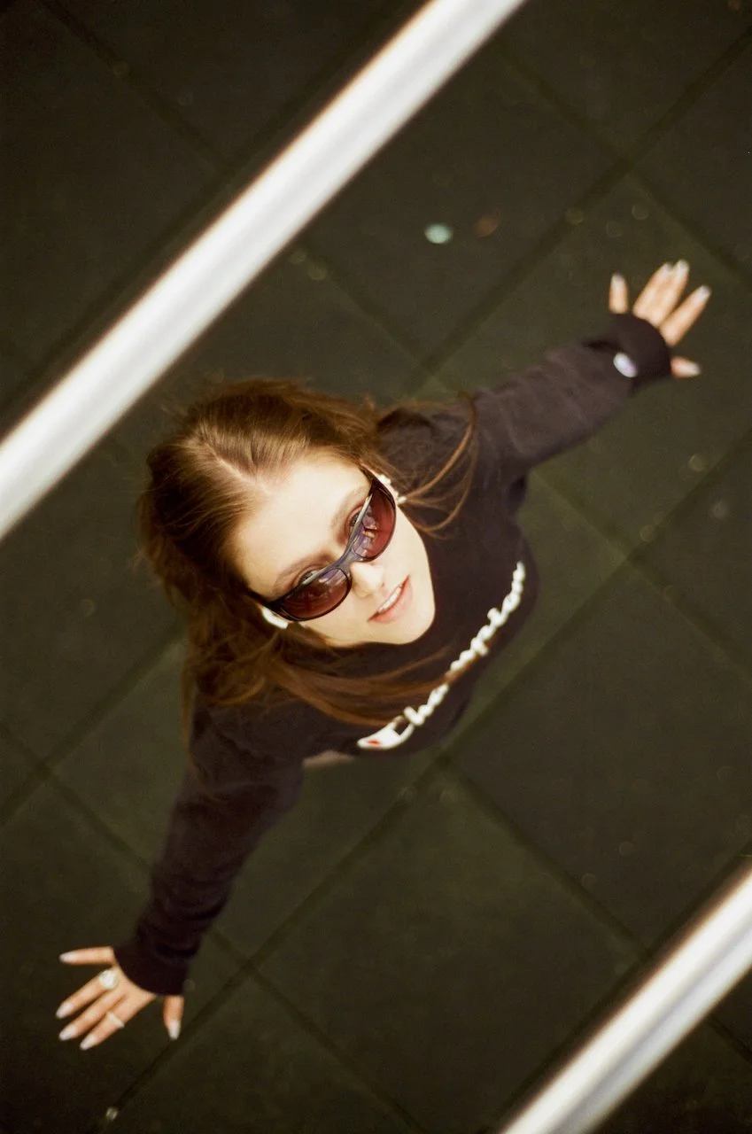 Junge Frau mit langen braunen Haaren, Sonnenbrille und schwarzer Jacke schaut nach oben, aufgenommen aus einer Vogelperspektive, während sie auf einem schwarzen Fliesenboden steht.