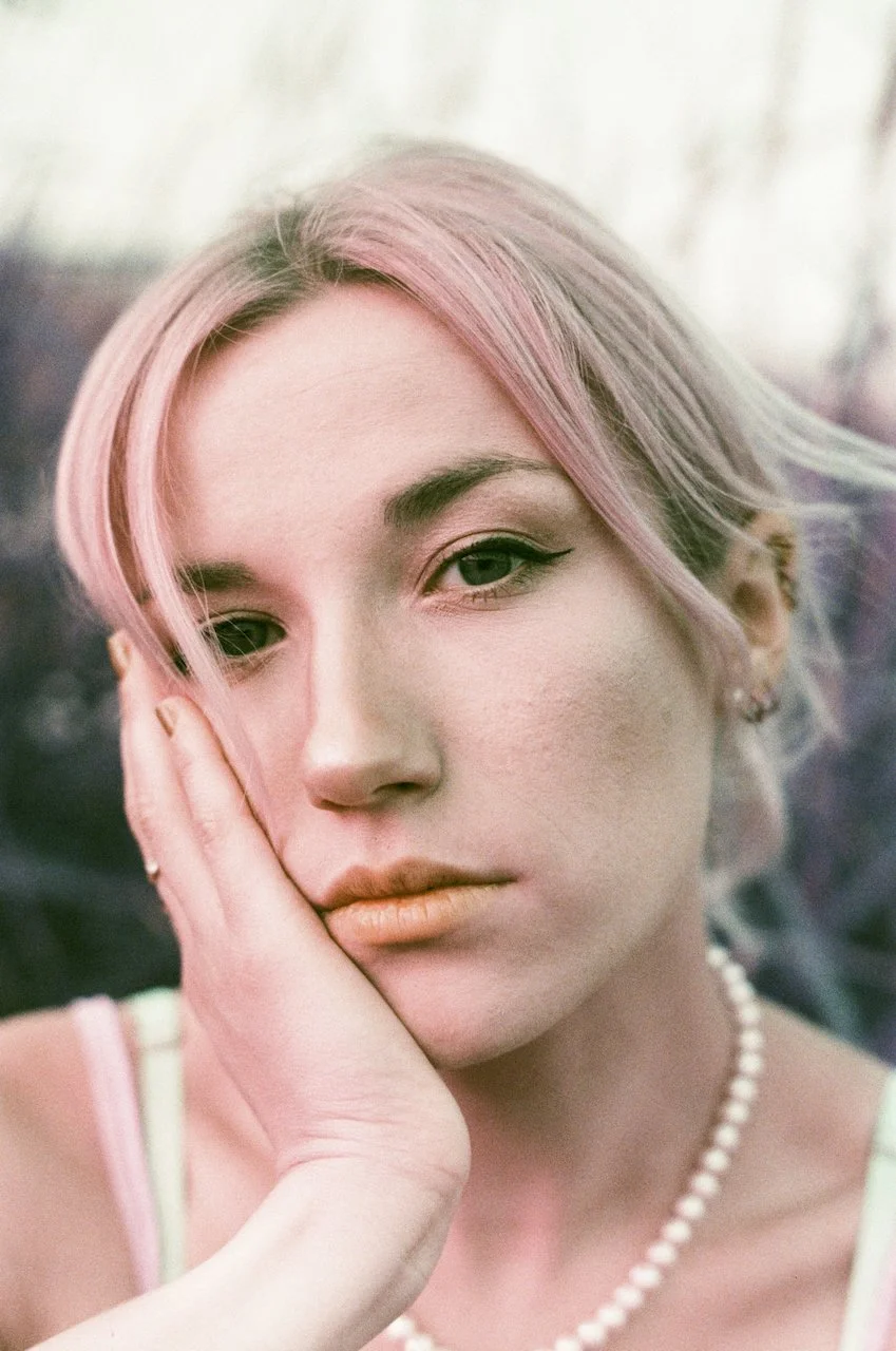 Nahaufnahme eines Mädchens mit rosa Haaren, das den Kopf auf die Hand stützt, trägt Perlenkette, make-up mit Eyeliner und Lippenstift.