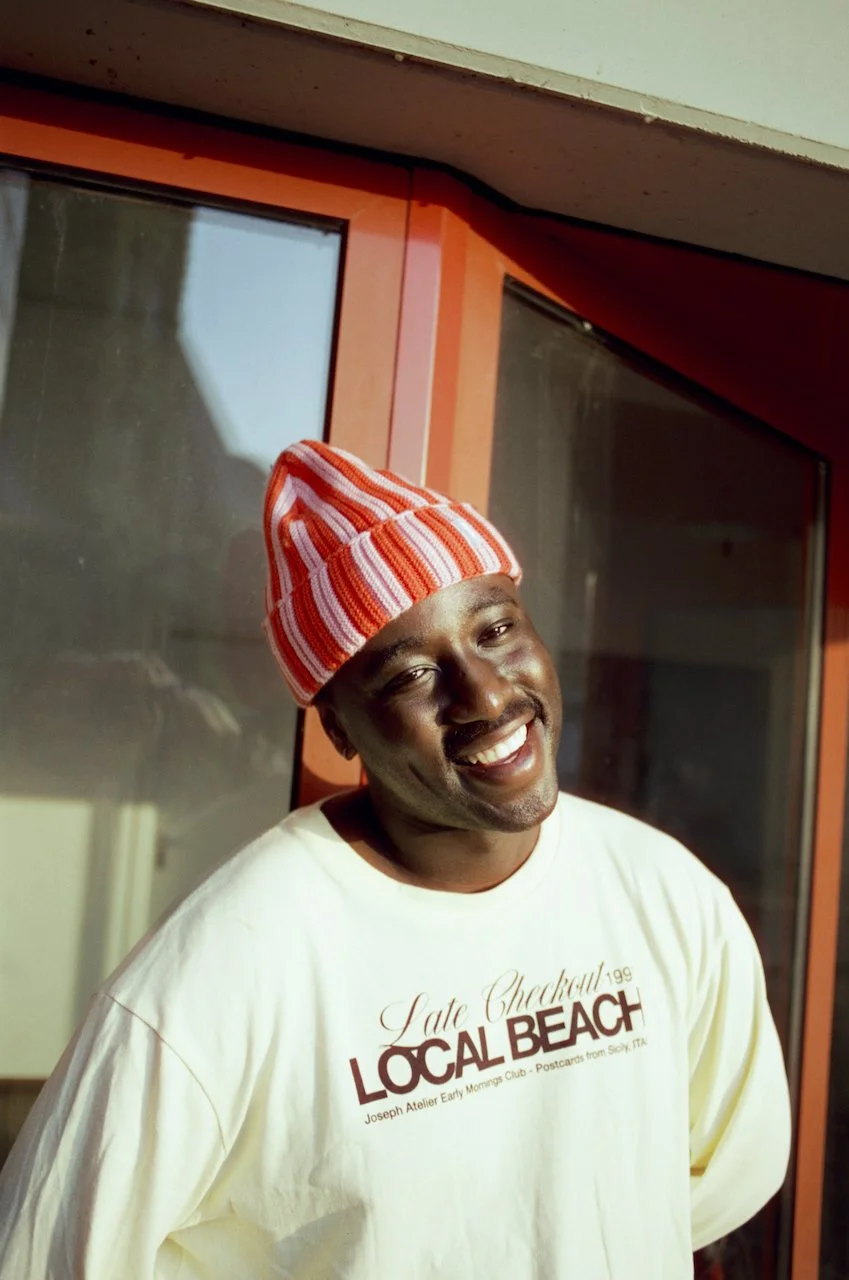 Ein lachender Mann trägt einen roten und weißen gestreiften Beanie und ein hellgelbes T-Shirt mit Aufdruck, im Hintergrund Fenster mit rotem Rahmen.