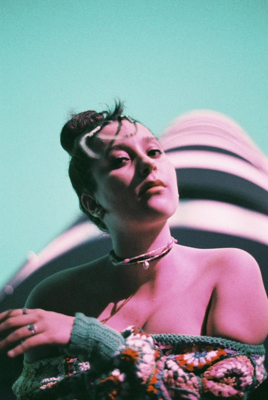 Porträt einer jungen Frau vor einem bunten, grafischen Hintergrund, mit bunten Haaren, Schmuck und einem bunten Strickpullover.