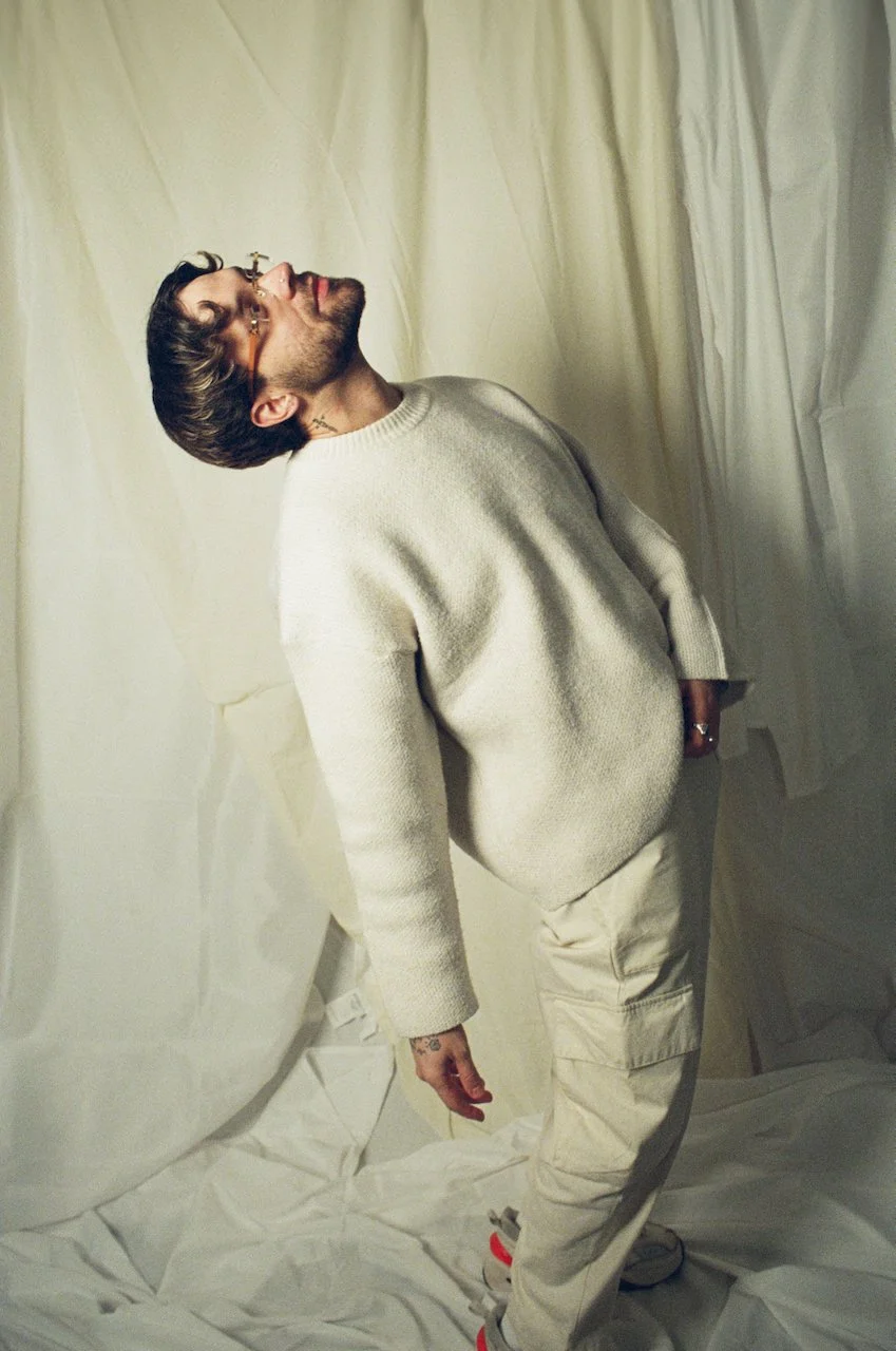 Ein Mann mit Brille, gestyltem Haar und einem Bart steht in einem hellen Raum vor einer beige Übergardine, trägt ein cremefarbenes Pullover und helle Hosen, schaut nach oben und hat eine entspannte Haltung.