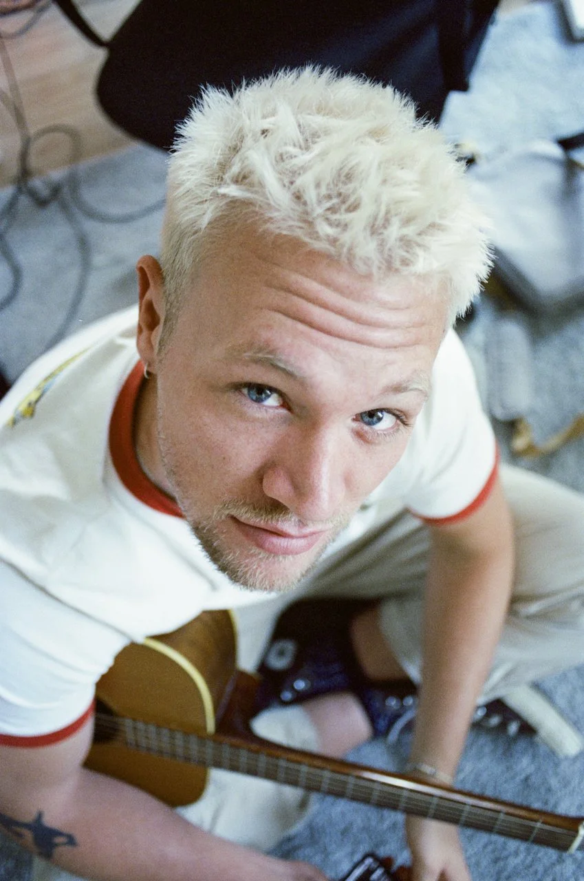 Junger Mann mit blondem, lockigem Haar sitzt mit einer Gitarre auf dem Boden, schaut in die Kamera und lächelt leicht.