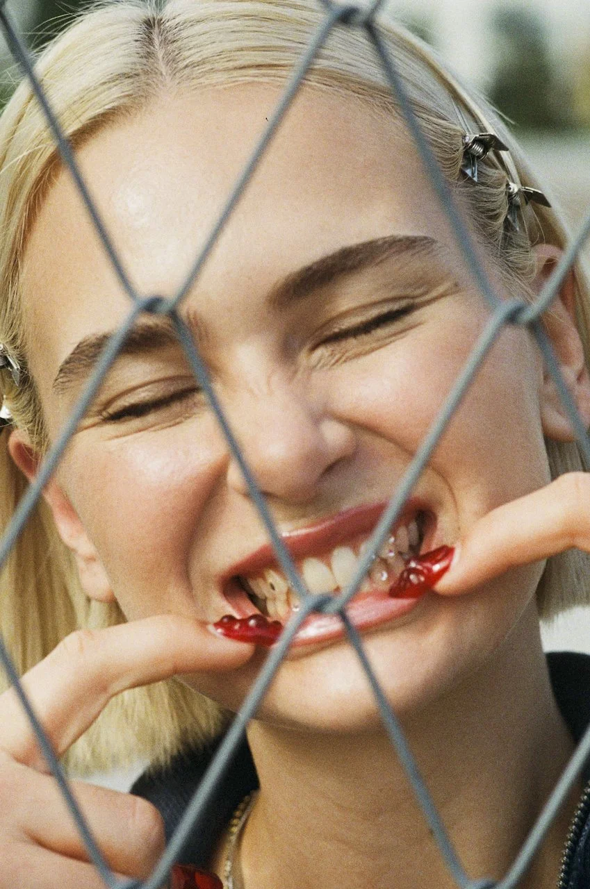Eine junge Frau mit blonden Haaren, die durch einen Gitterzaun schaut und an den Zähnen und Lippen spielt, während sie lächelt.